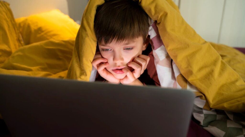 A young boy under a yellow blanket watching a laptop, appearing stress and focus in a dimly lit room.